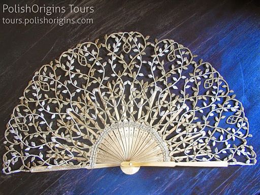 a detailed image of an antique, decorative lace fan. the fan features an ornate design with patterns of leaves and twigs. in the upper left corner is text: "polishorigins tours" and "tours.polishorigins.com". the fan is placed against a dark background, which is half black and half blue.