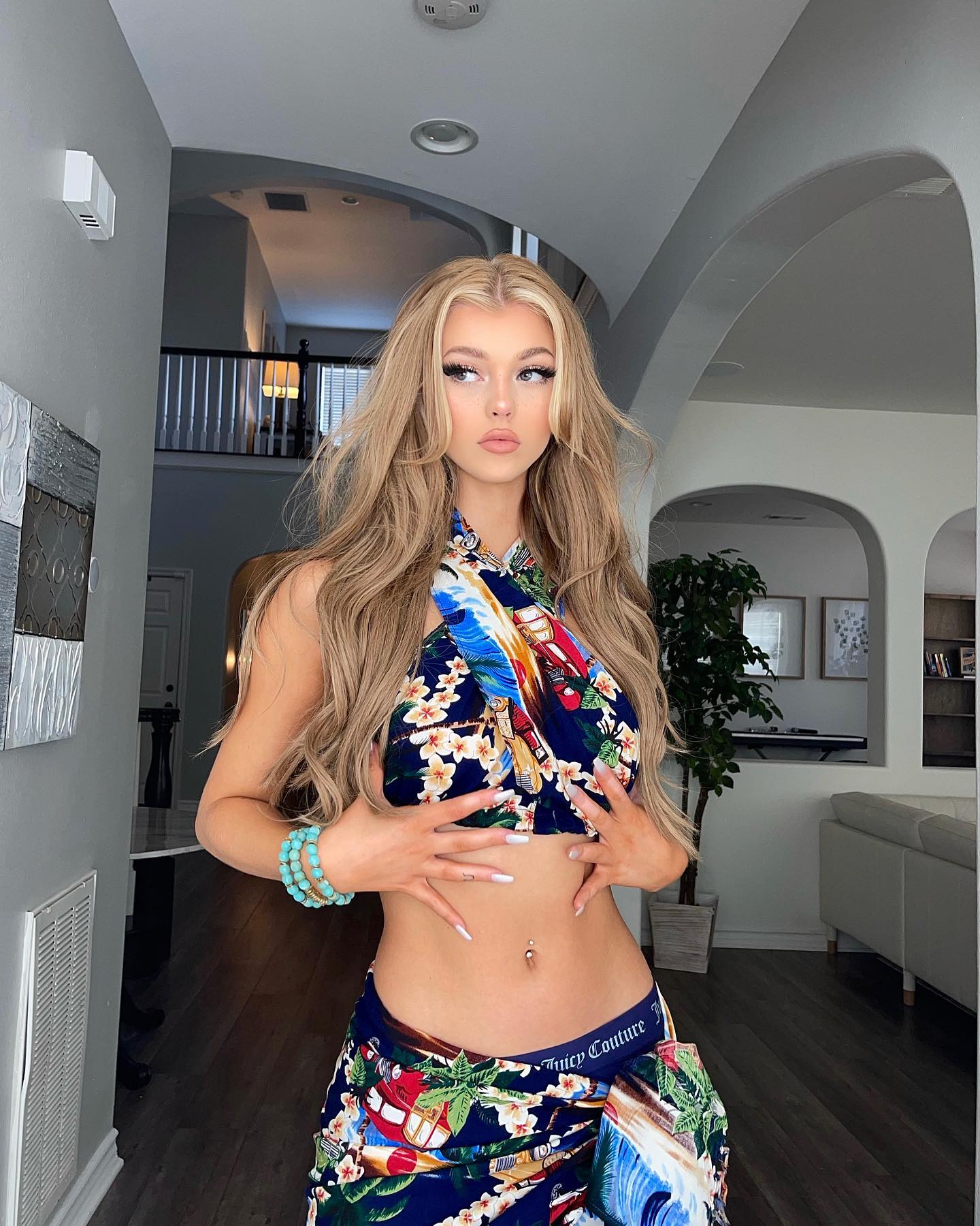 a young woman with blond hair is posing in a floral print halter top and skirt in a house interior. she is wearing a turquoise bracelet and has a navel piercing.