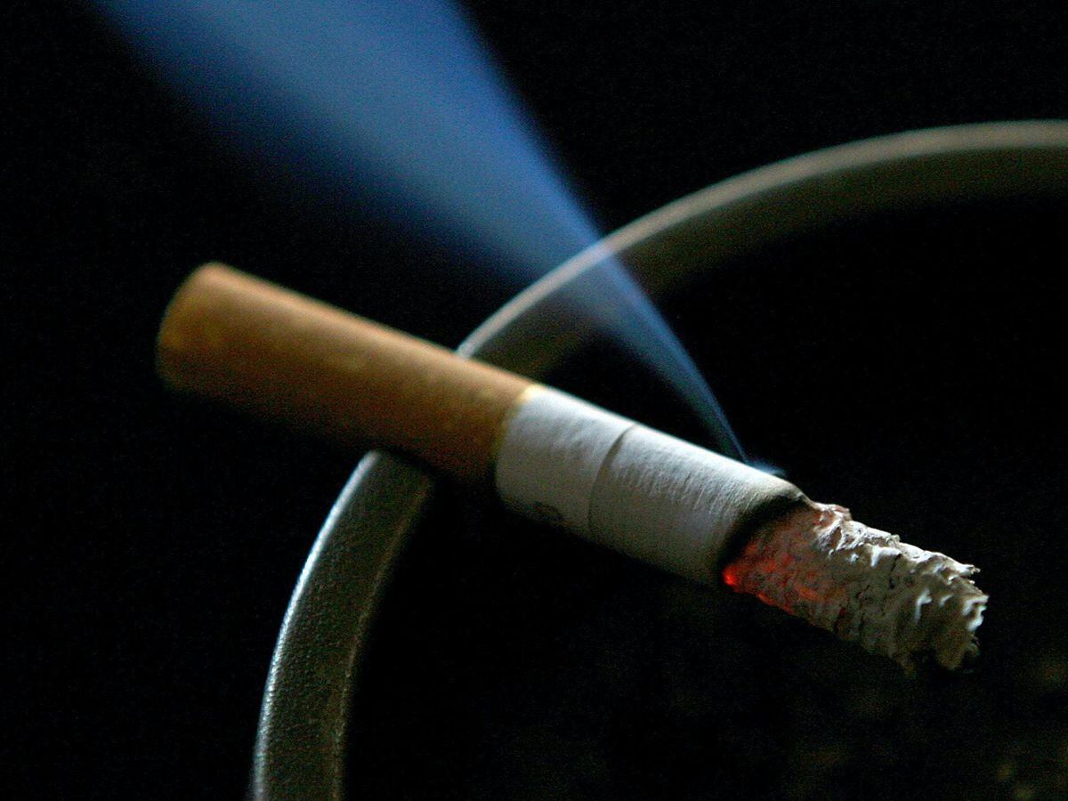 a lit cigarette rests in an ashtray, smoke curls upwards. the cigarette has a brown paper wrapper, a white filter, and burning ember with ash forming at the tip. the ashtray is metallic with a circular ring to hold the cigarette. the background is dark.