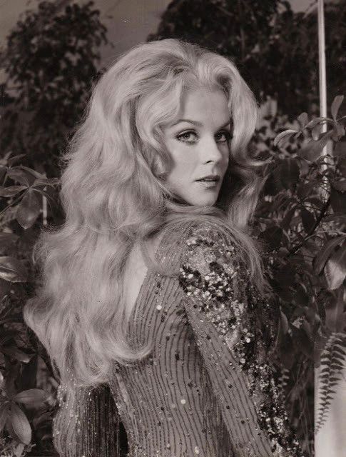 a glamorous young woman with long, blonde hair and a sequined dress looks over her shoulder at the camera. she is posing in a studio setting, perhaps for a magazine photo shoot. the image has a vintage feel.