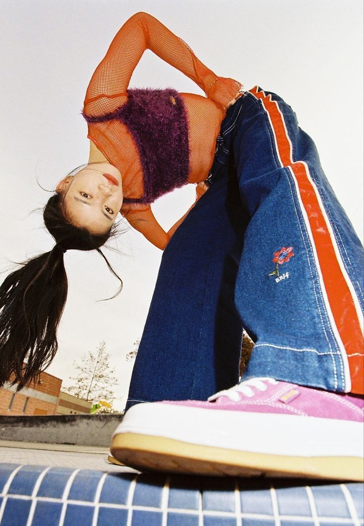 a young woman wearing a net orange shirt and jeans is standing in front of a skate park.  the image is taken from a low angle with the woman looking down at the camera. her long black hair is pulled back into a ponytail. she is wearing a pair of pink sneakers. she is smiling and seems to be having fun.