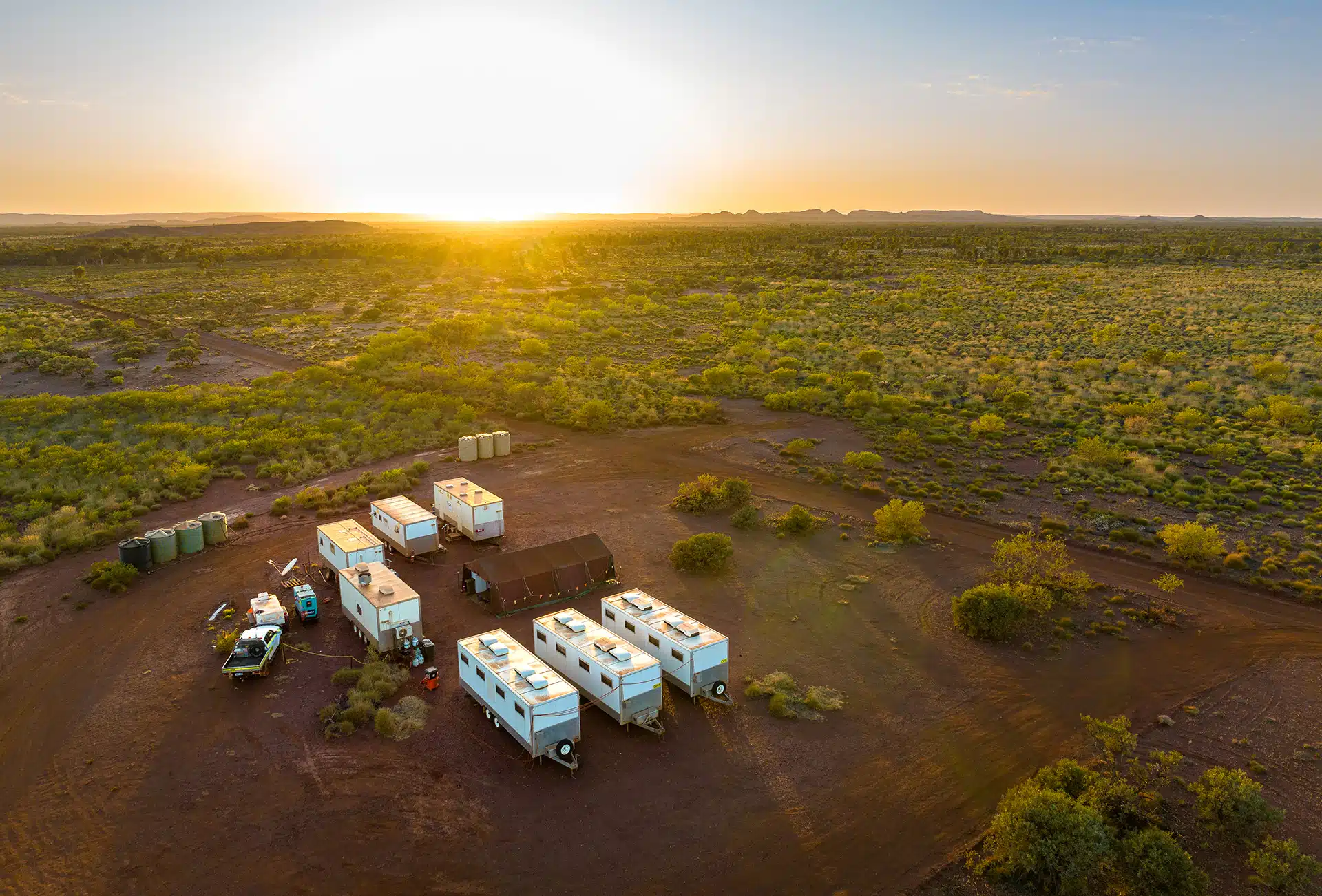 an aerial view of a remote campsite in the australian outback at sunset. the sun is setting in the distance, casting a warm glow over the landscape. there are several caravans and campers parked on the red dirt, and a few vehicles are visible. the scene is peaceful and serene, with the vastness of the desert stretching out in all directions.  the landscape is characterized by dry, arid conditions and sparse vegetation.