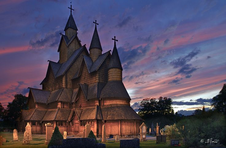 this image showcases a wooden church with intricate architecture, known as a stave church, set against a vibrant sunset sky. the sky displays hues of pink, purple, and blue, with scattered clouds. the church features multiple towers topped with crosses, and the surrounding landscape includes a graveyard with tombstones and crosses. trees are visible in the background, framing the church and contributing to the overall scenic view. the photo captures a serene and slightly ethereal atmosphere due to the lighting and composition.