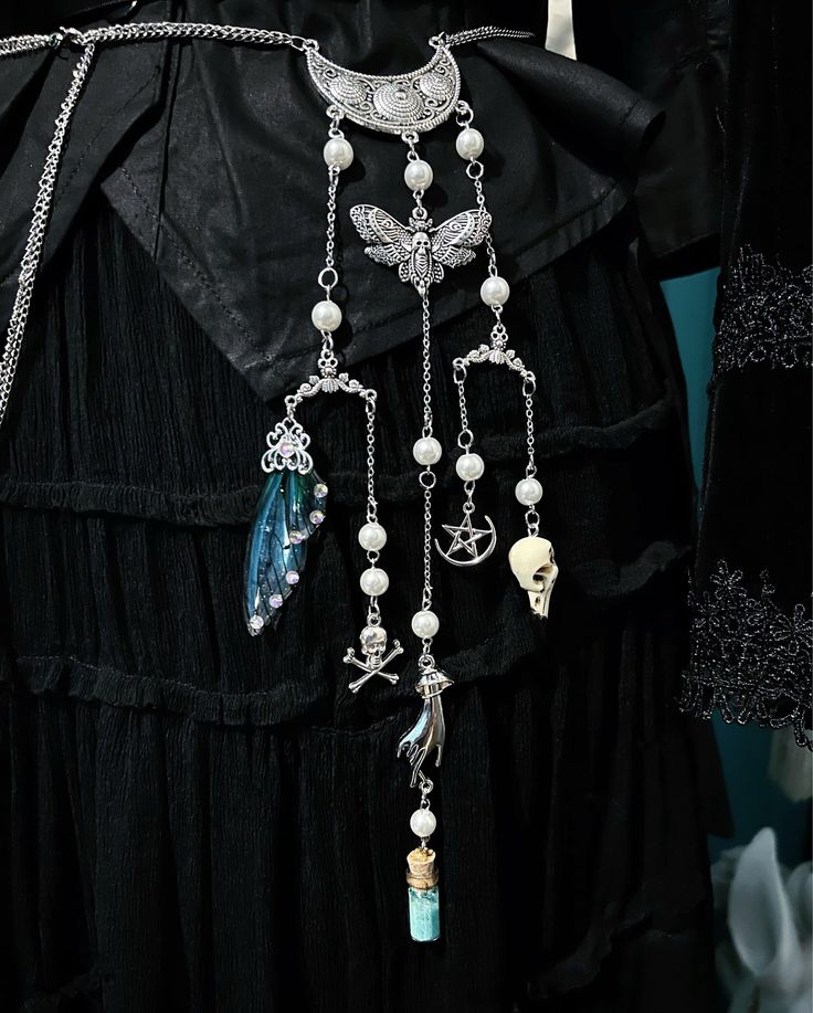 a closeup shot of a gothicstyle outfit featuring a black dress and a statement necklace. the necklace is made of a silver chain with various pendants, including a crescent moon, a butterfly, a skull, a hand, a potion bottle, a fairy wing, a pentagram, and a skull and crossbones. the necklace also features pearls. the dress is made of black fabric with ruffles and lace details.