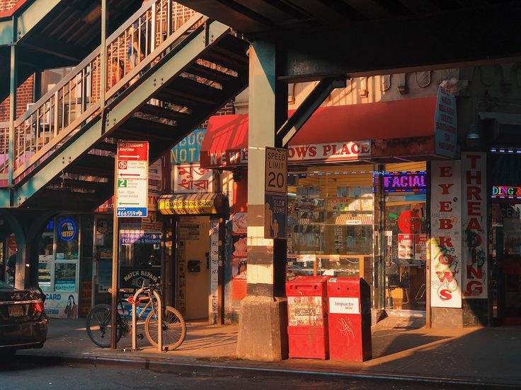 the image captures a quintessential new york city street scene bathed in the warm glow of sunset. an elevated train track casts long shadows across the street, highlighting a row of small businesses with colorful awnings and signs. a lone bicycle is chained to a pole, and a car is parked on the street. the warm light and long shadows create a sense of tranquility and nostalgia, offering a glimpse into the rhythm of city life.