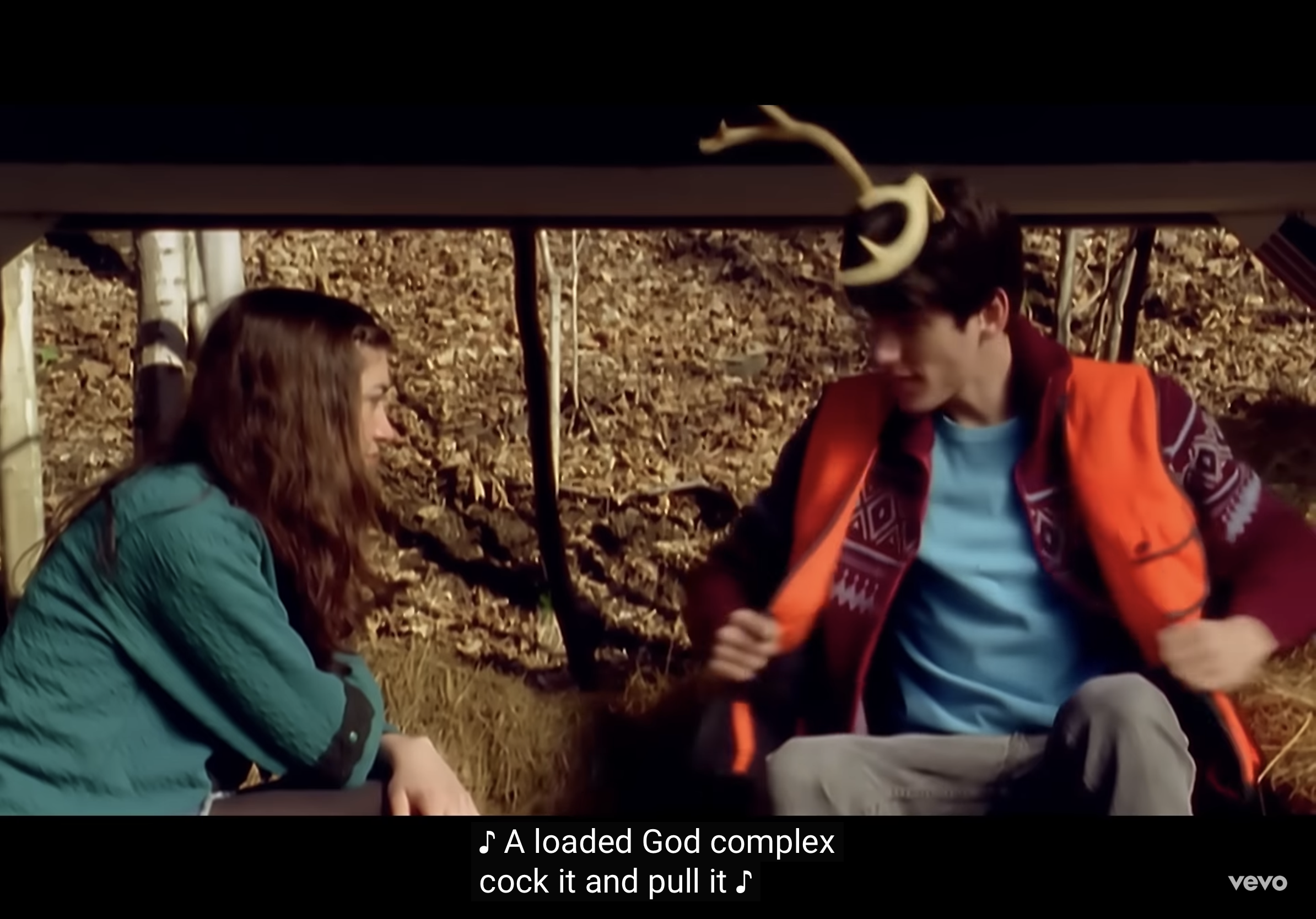 a still image from a music video depicting a young man and woman sitting beneath the open tailgate of a vehicle. the man, wearing a patterned vest, is in the process of removing or adjusting his outer layer, while the woman with long brown hair looks at him intently. the setting is a forest with fallen leaves on the ground, bathed in soft, natural light that suggests daytime. the overall mood is intimate