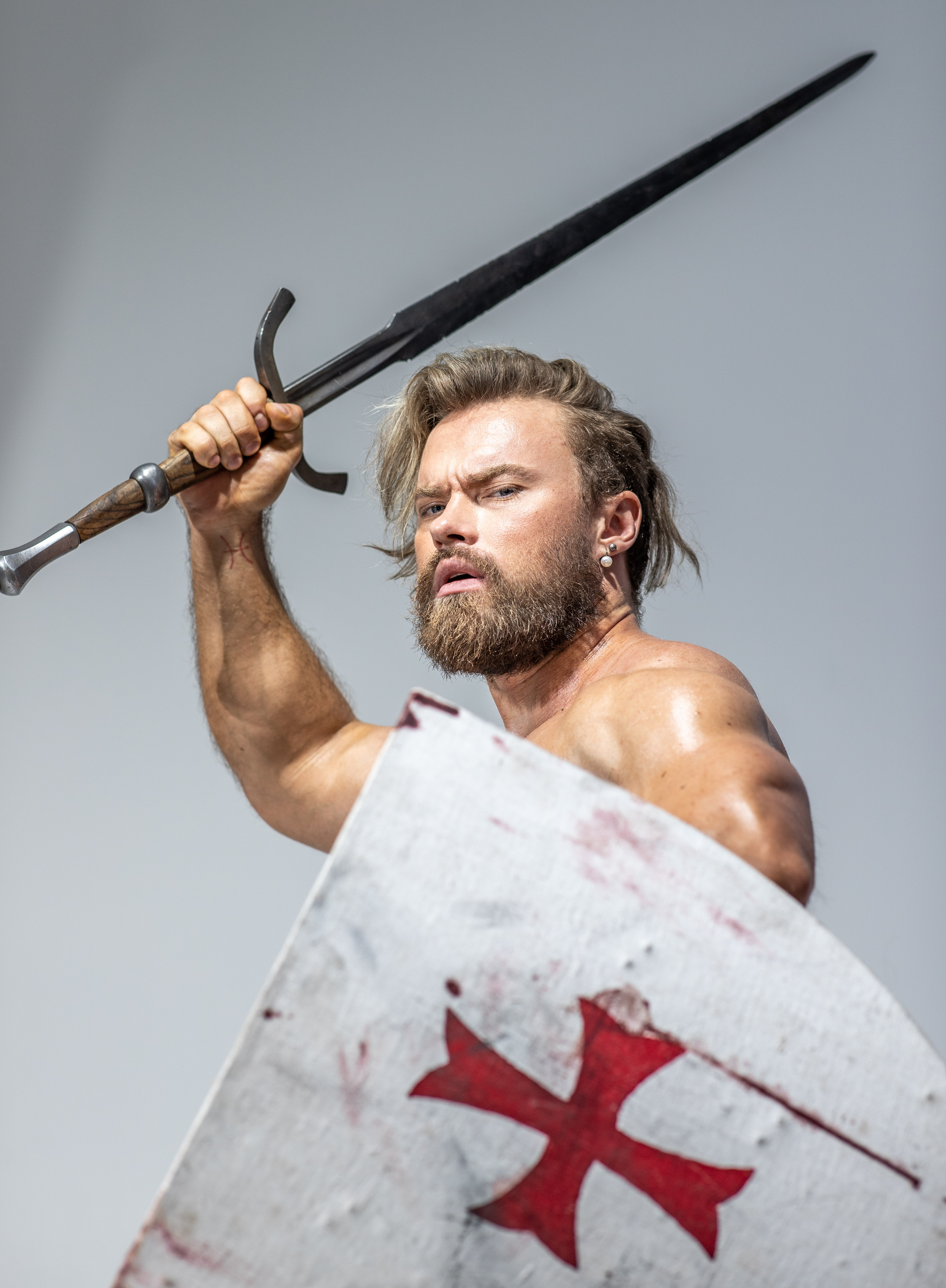 a muscular man with a beard, styled as a medieval warrior, dominates the frame. he's shirtless, showcasing his physique, and wields a gleaming sword raised high above his head. he grips a large, round shield, emblazoned with a bloodred templar cross, partially obscuring his lower body. the shield bears the scars of battle, hinting at his fierce nature. the studio lighting casts a heroic glow upon him, emphasizing his strength and determination.  his intense gaze focuses forward, suggesting he's in the heat of battle.