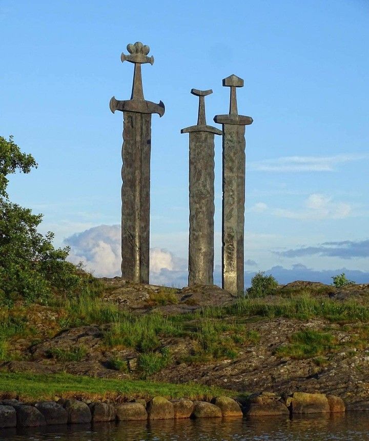 the image shows the sverd i fjell monument, also known as swords in rock, located in hafrsfjord, stavanger, norway. three large viking swords are planted in the rock, commemorating the historic battle of hafrsfjord in 872 ad which unified norway. the monument is set against a backdrop of a blue sky with scattered clouds, with a rocky landscape leading down to the water's edge. trees are visible on the left side of the image.