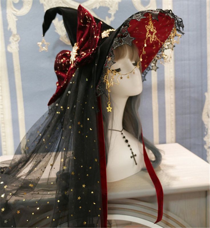 this is a closeup shot of an elaborate witch hat with a celestial theme. the hat is black with a wide brim, adorned with gold stars and lace. a large velvet bow in deep red with gold star accents sits on the front, complementing a red underbrim. a long black veil speckled with gold stars drapes from the hat, partially obscuring the mannequin head wearing it. the background is kept simple and bright to highlight the details of the hat.