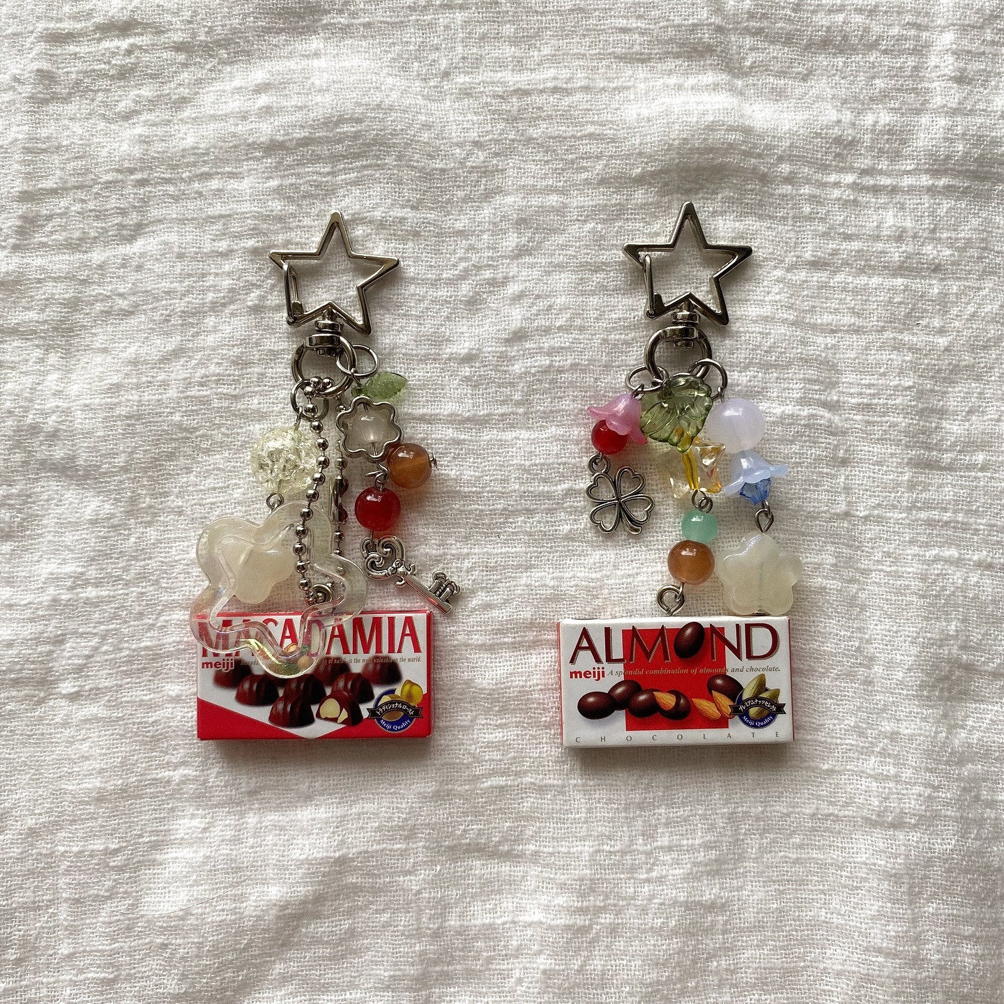 the image shows two handmade keychains featuring candy box trinkets hanging from starshaped clips, adorned with various beads, charms, and trinkets. on the left is a keychain featuring a meiji macadamia chocolate box, with a clear unicorn, flowers, beads, a chain, and a tiny key dangling above. on the right, the keychain features a meiji almond chocolate box, with a metal clover, floral beads, and other colorful trinkets.
