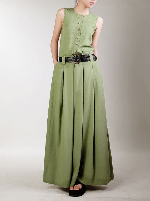 a woman is shown wearing a long, sleeveless, green dress. the dress features a buttondown top and a belted waist with a woven belt and a round buckle. the dress has pockets and pleats from the waist down, creating a full skirt. the woman is wearing black sandals. the background is a light, neutral color.