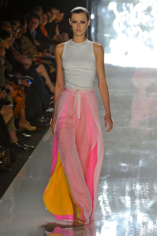 a model walks down a runway wearing a white sleeveless top and pink and yellow chiffon pants. the audience is blurred in the background.