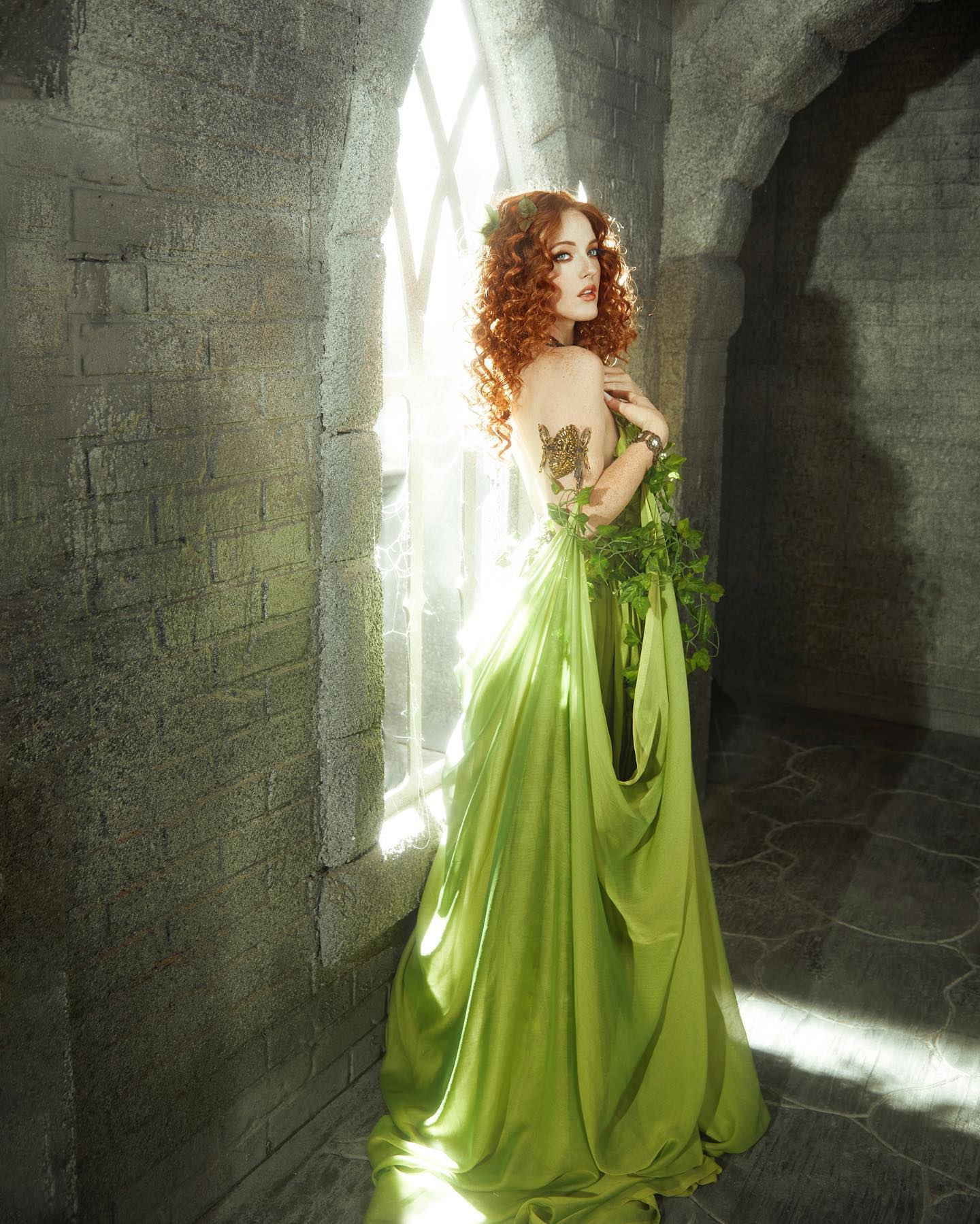 a woman with long, flowing red hair stands in the light streaming through a castle window. she's wearing a flowing green gown with ivy accents, evoking a sense of fantasy and nature. the setting and her attire suggest she could be a mythical queen or goddess.