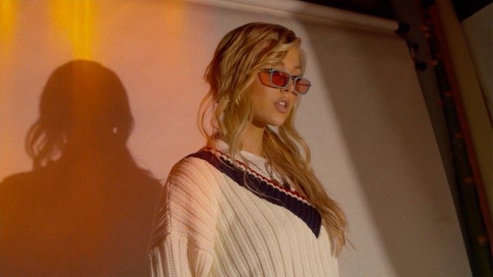 a medium shot shows a blonde woman standing indoors, wearing a white sweater with blue and red trim and tinted glasses. an orange light casts a shadow of her figure on the backdrop. the scene appears to be in a studio setting.