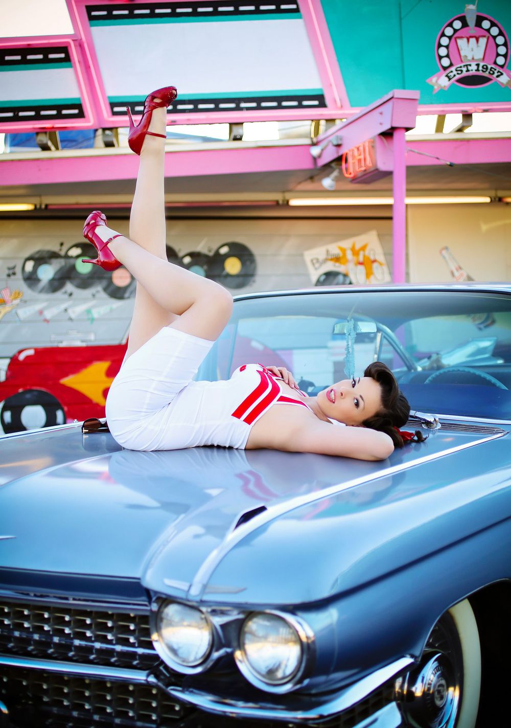 a pinup girl in a white dress with red accents lies on the hood of a vintage light blue cadillac. she's wearing red high heels and her legs are up in the air. the background features a retrostyle diner with pink and teal colors. the scene evokes a classic 1950s aesthetic. the image is bright and welllit, showcasing the model's beauty and the car's classic lines.