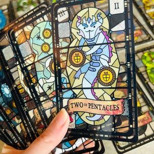 a hand holds a stack of tarot cards. the top card is the "two of pentacles" depicted in a stained glass style, featuring a light blue dragonlike creature balancing two pentacles. the cards have a black frame and showcase various suits in a similar stained glass design.