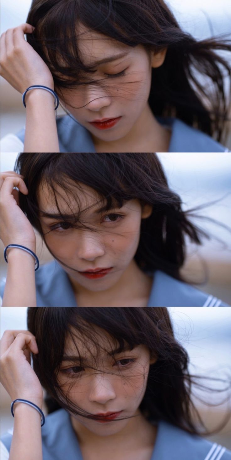 this image consists of three slightly different closeup portraits of a young asian woman with dark hair blowing in the wind. she is wearing a blue garment and has a simple blue bracelet on her wrist. her expression is thoughtful, perhaps melancholic, and the overall aesthetic is soft and natural. the setting appears to be near a beach or coastal area.