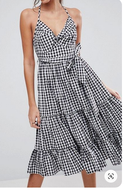 a woman models a black and white gingham midi dress. the dress features a halter neckline, a tie waist, and a tiered skirt. the image is taken from the front and shows the dress from the waist up. the background is white.