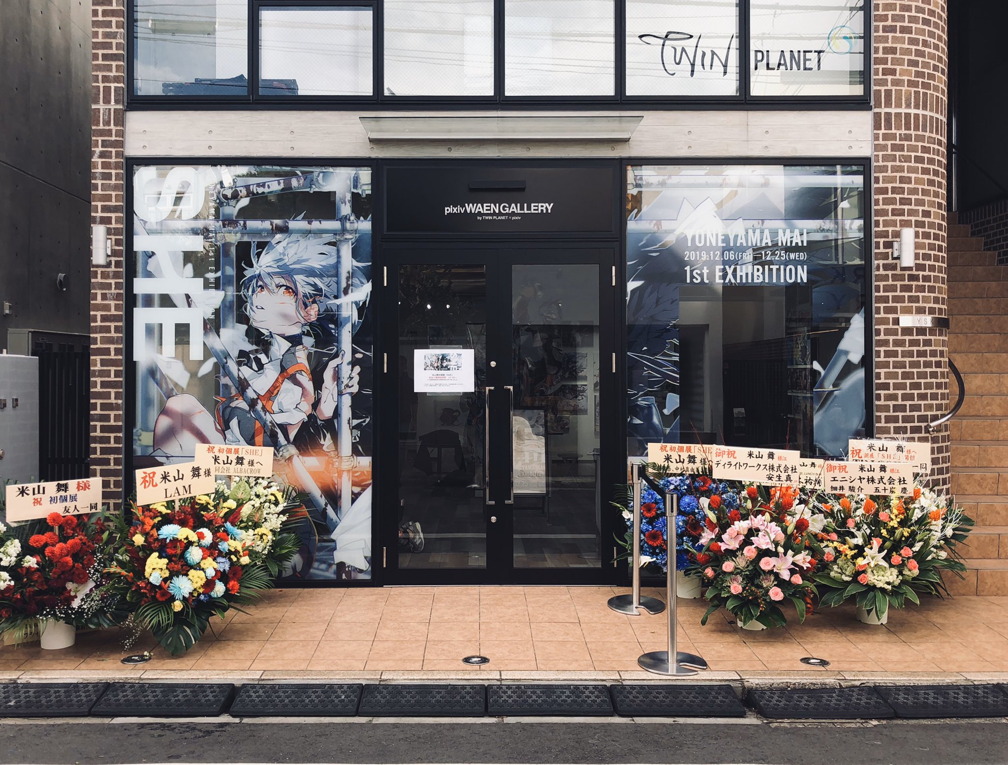 a photo of the storefront of an art gallery with a large anime graphic on the window. there are bouquets of flowers arranged on either side of the door. the sign above the door says "twin planet" and "photo waengallery". there is a banner on the right side of the door that reads "tunetama mai 2019.12.0612.25 1st exhibition".