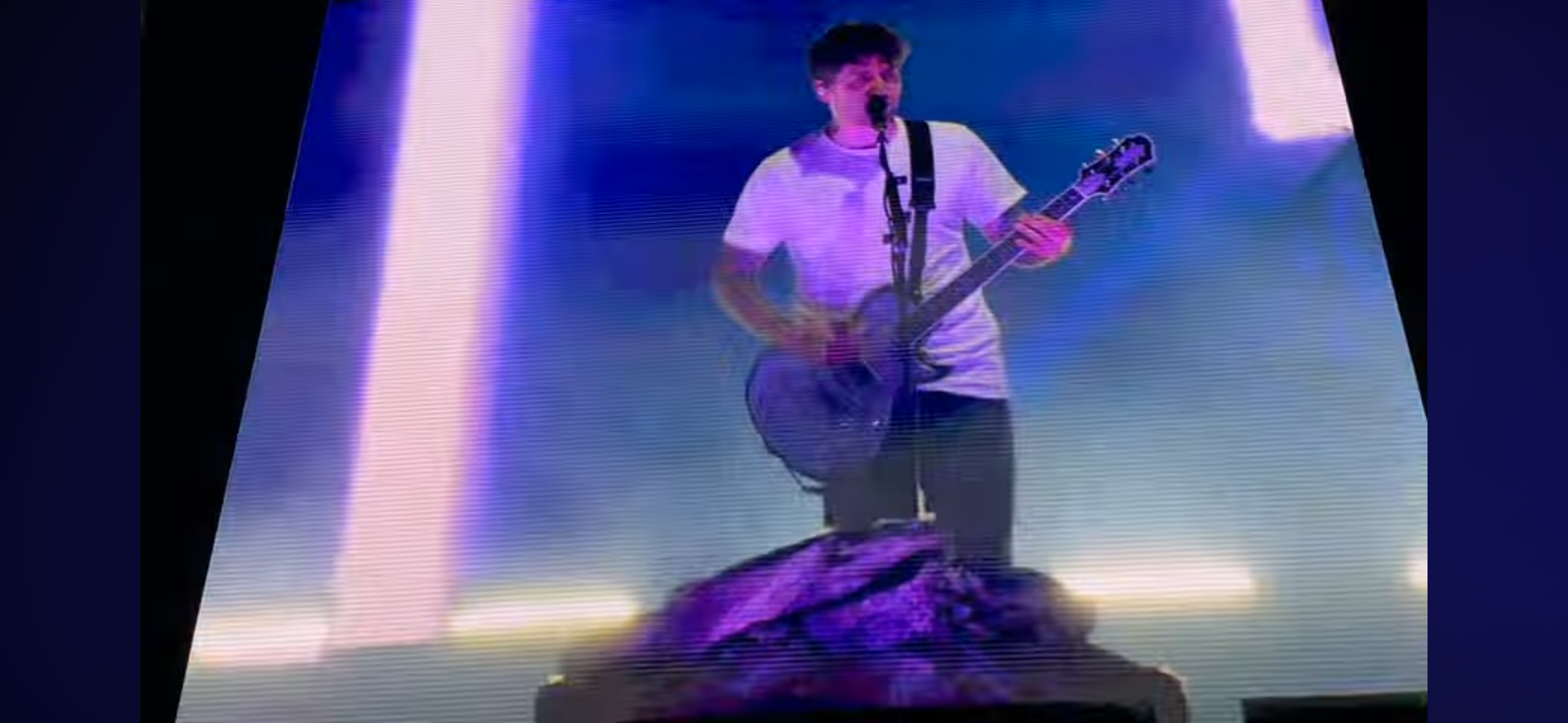 a man in a white shirt and black pants plays an electric guitar on a stage with purple lights. he's singing into a microphone, and there's a crowd of people cheering in the background. he's giving a great performance.