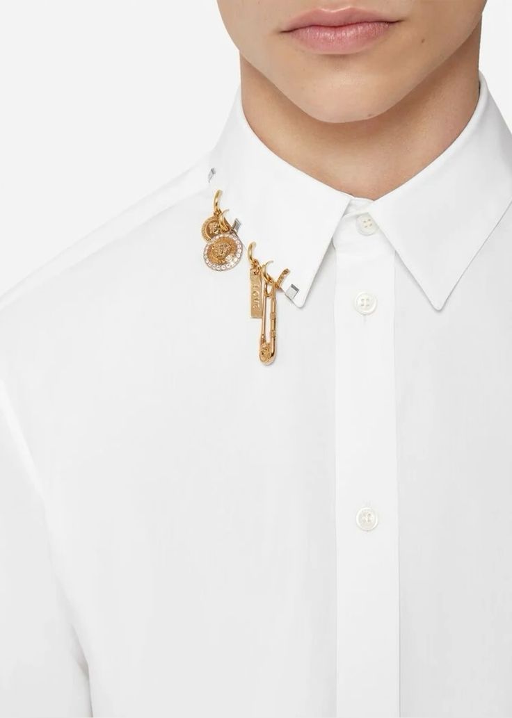 the image shows a person wearing a white buttondown shirt. the shirt has a collar with gold jewelry attached to it. the jewelry consists of a gold pin and other gold ornaments. the brand of the jewelry and the shirt appears to be versace.