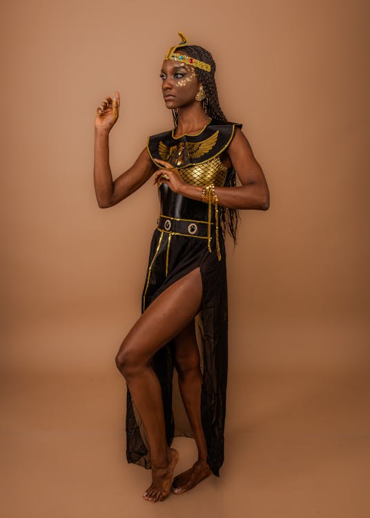 a woman dressed in an elaborate egyptian costume poses against a solid brown background. she is wearing a black and gold dress with a high side slit, a matching headdress, and gold jewelry. her dark skin contrasts with the bright gold accents of her outfit and accessories. she has braids in her hair and dramatic makeup. she is barefoot and in a dynamic pose, with one hand raised and the other gesturing towards her chest. the photo is a studio shot, highlighting the costume and the model's presentation.