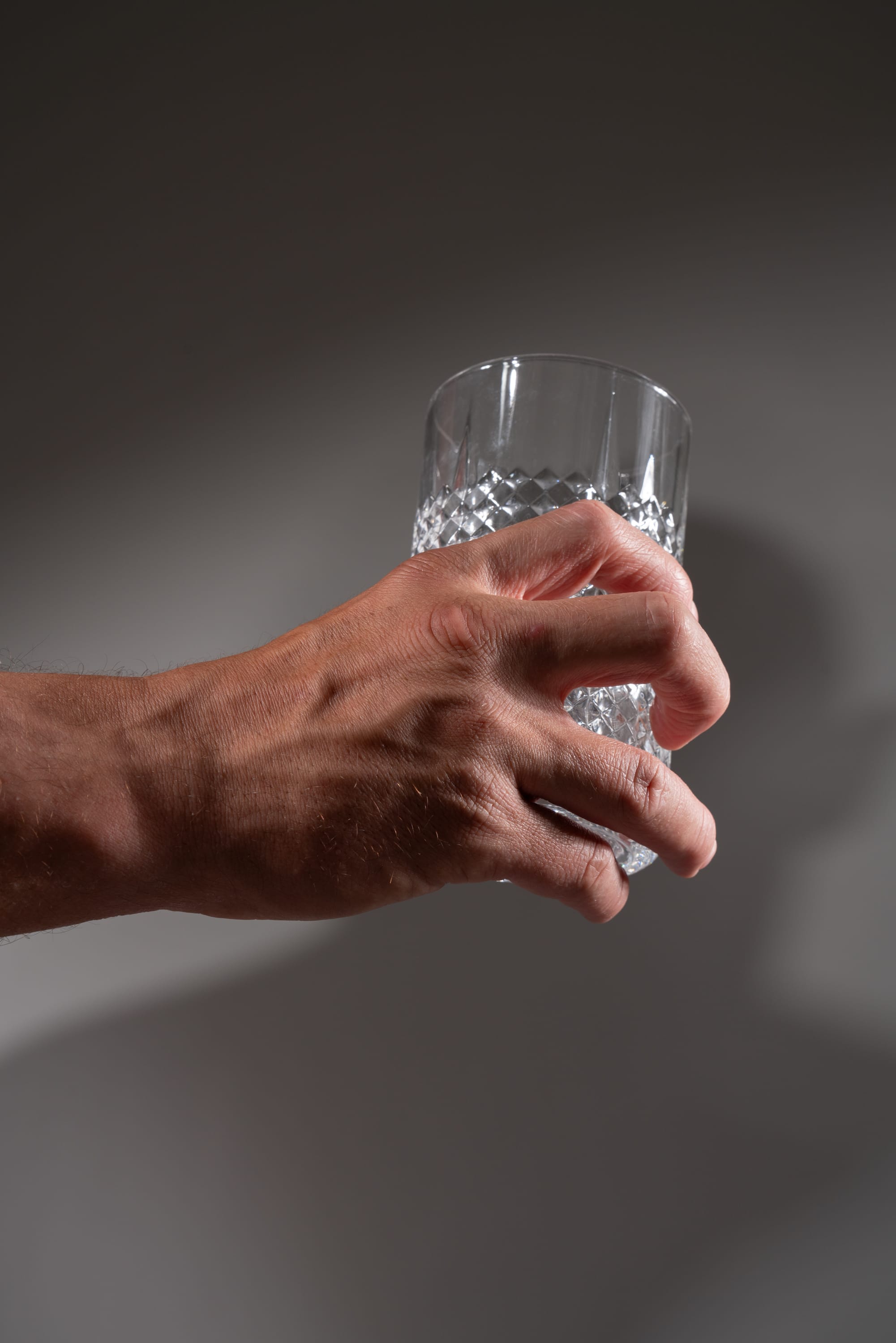 a closeup image of a man's hand holding an empty, clear cut glass against a gray background. the studio lighting creates a distinct shadow of the hand and glass on the background.