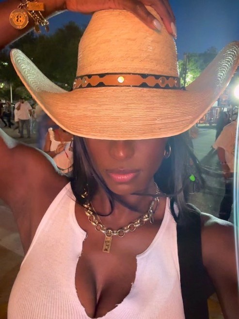 a woman is smiling as she wears a straw cowboy hat, white tank top, and gold necklace while looking directly at the camera. she is outdoors in the nighttime.