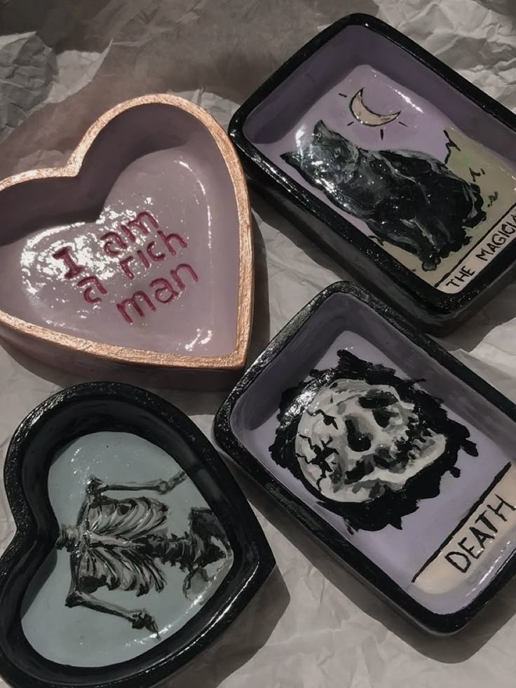 the image shows four trinket dishes arranged on a wrinkled white surface. one trinket dish is heartshaped and pink, with the phrase "i am a rich man" written inside in pink letters. another heartshaped trinket dish is black with a white skeleton design inside. the other two trinket dishes are rectangular, with one depicting a black cat and a crescent moon, labeled "the magician," and the other showing a skull and a cracked white moon, labeled "death."