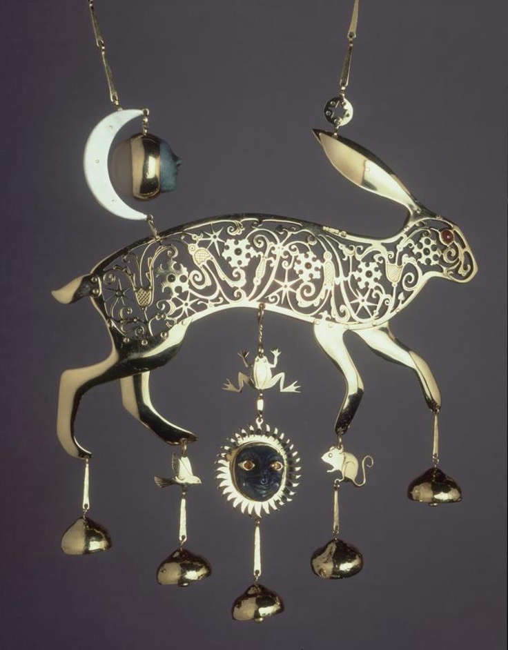an elaborate gold mobile features a hare in profile, suspended with various elements. the hare's body is intricately cut out with swirling designs, and a red stone marks its eye. hanging below the hare are small pendants: a frog, a sun with a face, a mouse, and three teardropshaped ornaments. above the hare, a crescent moon and a sculpted face dangle from chains. the entire piece is suspended against a plain background, highlighting its intricate design.