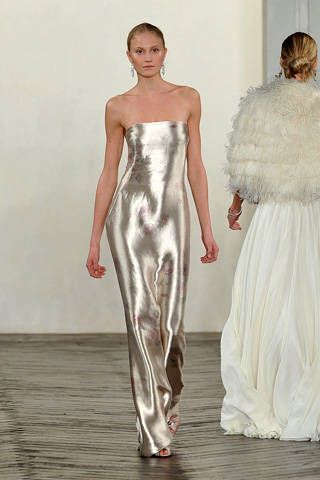 the image shows two women on a runway at a fashion show. the woman in the foreground is wearing a long, strapless silver dress. the woman in the background is wearing a white dress with a fluffy, featherlike bolero. both women are walking towards the viewer.