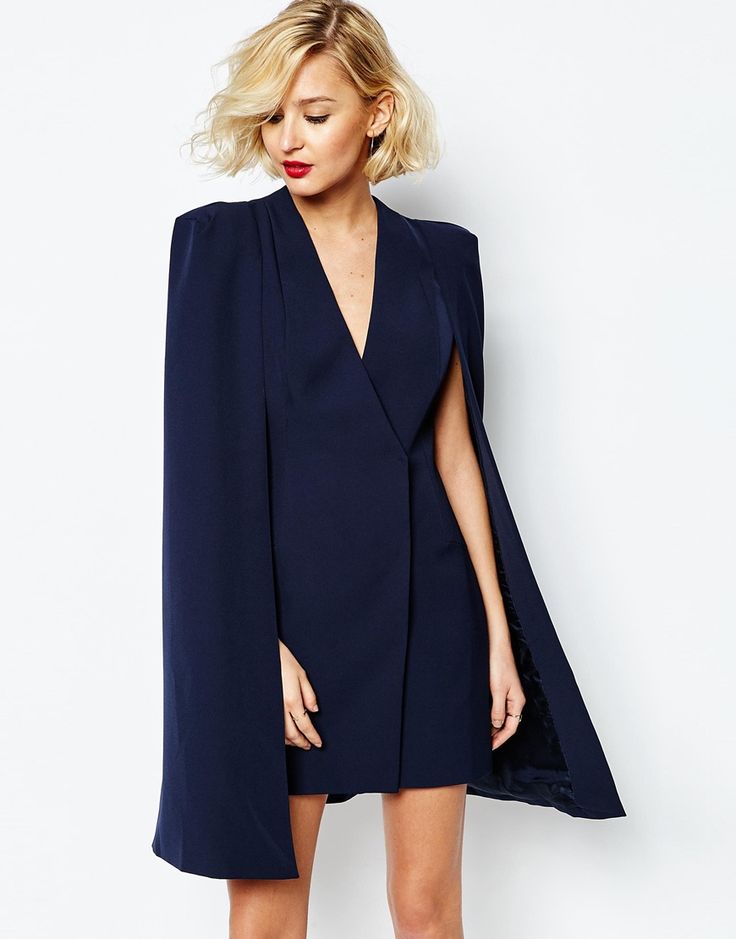 the image shows a fashion model, a blonde woman with shoulderlength wavy hair, wearing a dark navy blue dress. the dress is a unique design, featuring a capelike overlay that extends from the shoulders to below the knees. the dress is fitted at the waist and above the knee in length. it has a vneck and a  wrapstyle closure. the model is posing against a plain white background, looking slightly down. the overall style suggests a formal or semiformal occasion.  the image is professionally taken, with a focus on the clothing and its details.