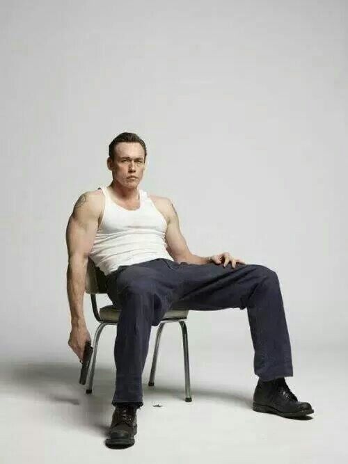 a man is sitting in a chair in a studio setting. he is wearing a white tank top, black pants, and black shoes. he is holding a gun and has a serious expression on his face. the background is a white wall.