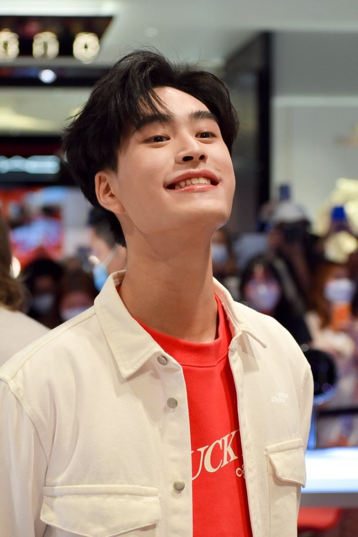 a portrait of a young asian man with short black hair wearing a white jacket and a red shirt. he is smiling brightly at something offcamera. the background is slightly blurred and suggests an indoor public space with other people present. he appears to be at a fansign event.