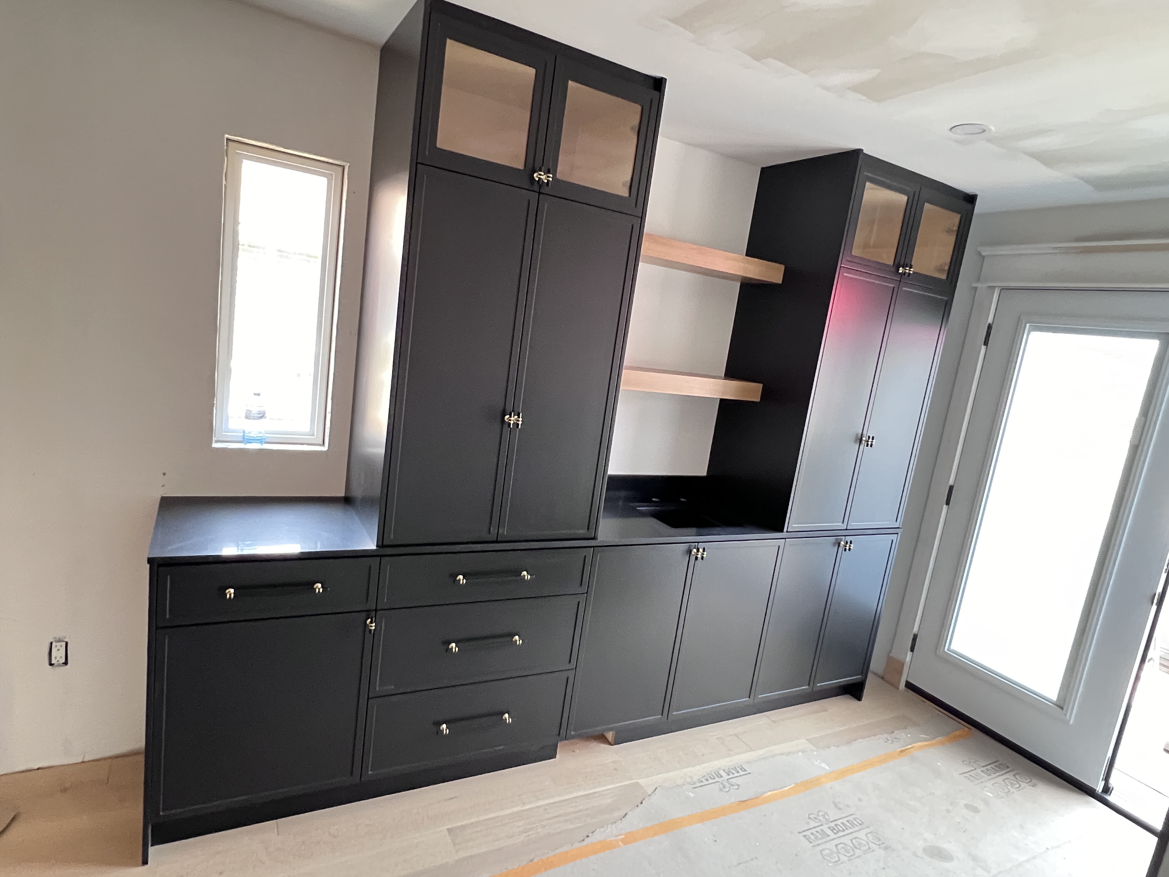the image shows a newly installed darkgray custom kitchen cabinetry with gold hardware. the cabinetry includes upper cabinets with glass doors, lower cabinets with drawers, and floating wooden shelves. the countertop is darkcolored and appears to be quartz. the room is under construction and includes hardwood flooring and a new french door.  there is also a window visible on the left. the overall style is modern and contemporary.