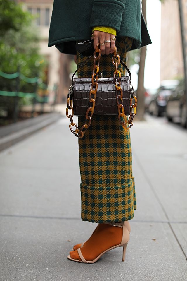a woman wearing a green coat, gingham pants in green and yellow, orange socks, and beige ankle strap heels is walking down a city street. she carries a brown crocodiletextured handbag with a chunky tortoise shell chain strap. the overall aesthetic is fall fashion with autumnal colors and textures.