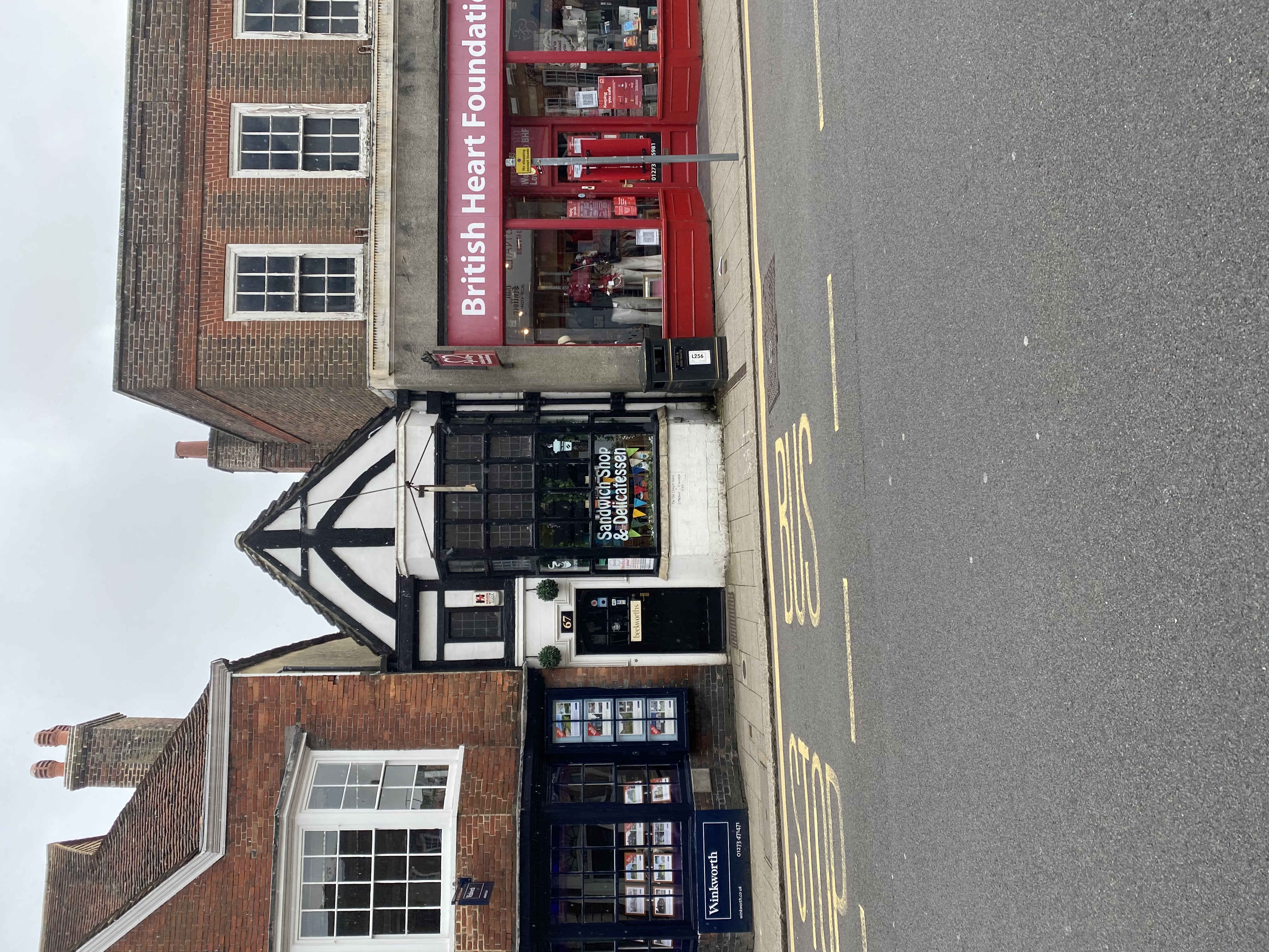 a photo of two buildings on the street. the building on the left is a brick building with a storefront that says "sandwich shop & delicatessen".  the building on the right is a brick building with a storefront that says "british heart foundation". there is a bus stop in front of the buildings.