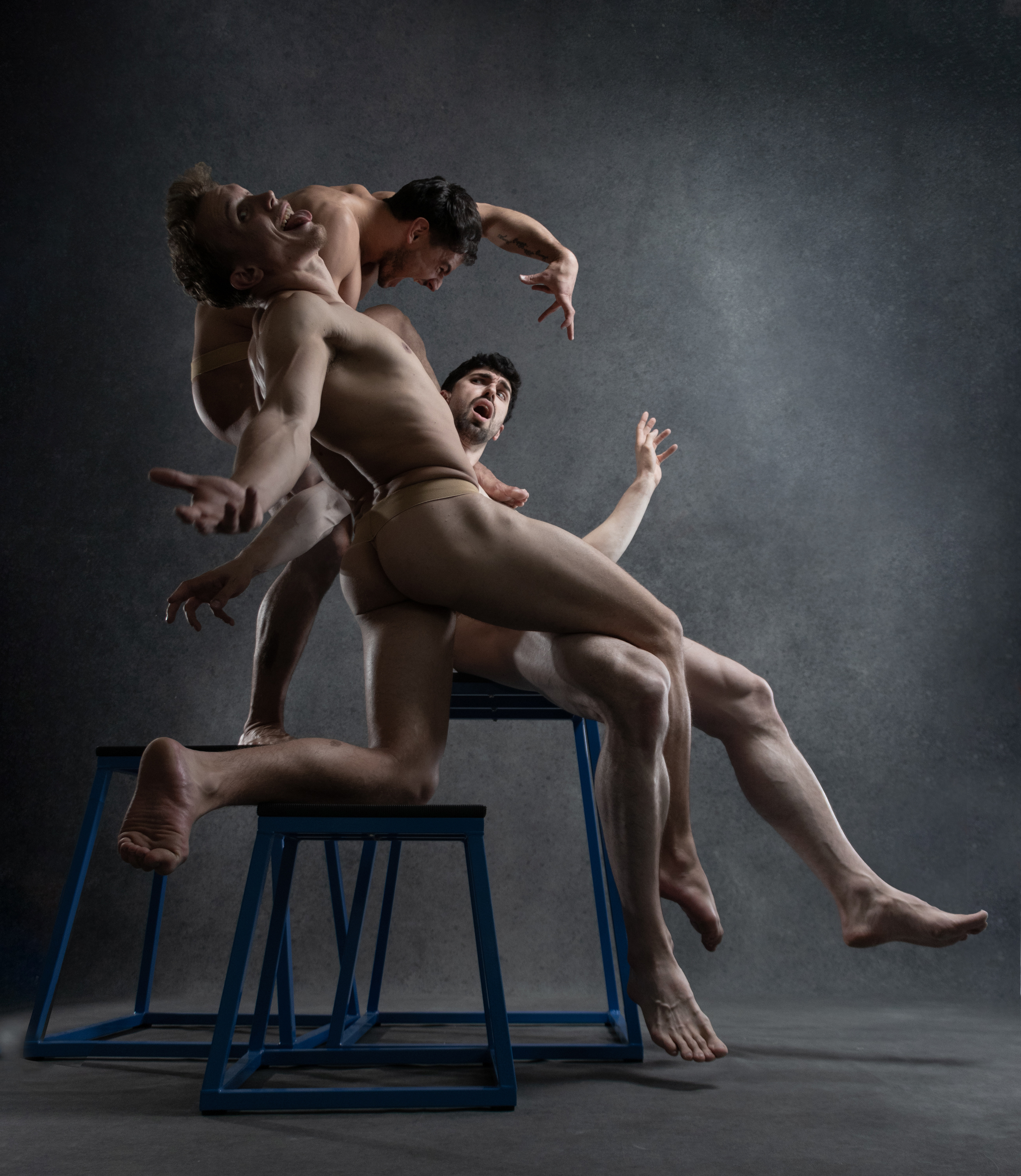 four muscular men are captured midair in a dynamic and expressive dance pose. they are shirtless and wearing only dance briefs, highlighting their toned physiques.  they are intertwined and balanced on a series of three blue metal stools, creating a sense of weightlessness and defying gravity. their faces are contorted with emotion, showcasing the intensity of their performance. the backdrop is a dark gray, providing a stark contrast to the dancers' bodies and adding to the dramatic atmosphere of the image.