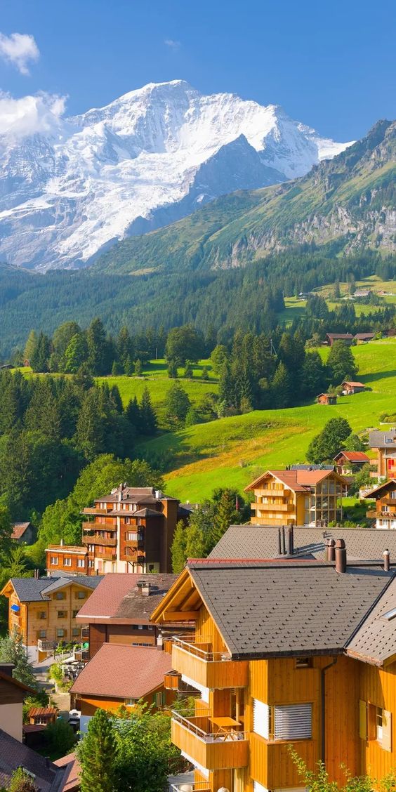 the image shows a picturesque swiss village nestled in a valley, with the majestic swiss alps towering in the background. the snowcapped peaks of the mountains are illuminated by the sun, creating a stunning contrast against the clear blue sky. the lush green slopes of the valley are dotted with charming wooden houses, their rooftops glistening in the sunlight.  the overall scene evokes a sense of tranquility, beauty, and the allure of the swiss alps.