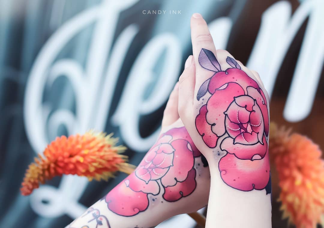 a closeup photograph showcases two hands adorned with vibrant neotraditional tattoos of pink flowers.  the tattoos, inked on the back of each hand and wrist, boast a captivating blend of pink hues with subtle hints of green and gray in the leaves.  the image appears to be taken inside a tattoo shop, evidenced by the blurred signage in the background.  the lighting is bright and seemingly natural, illuminating the intricate details and color saturation of the tattoos.