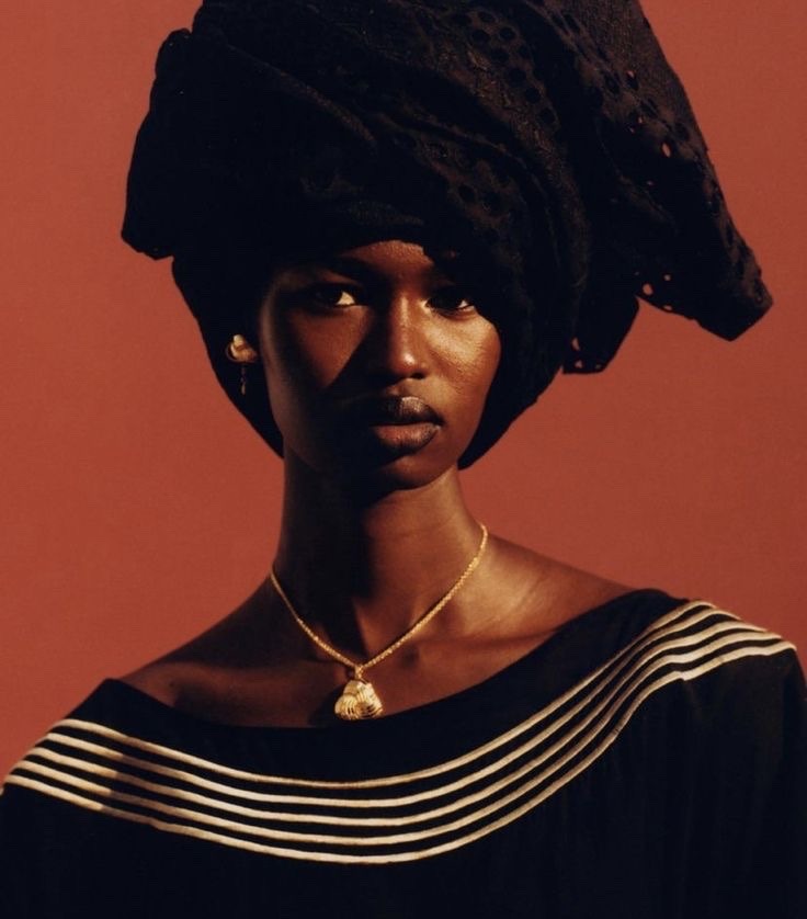 a portrait of a black woman wearing a head scarf and a gold necklace against a red background. the woman's skin is a rich brown color and her expression is serious. she looks directly at the camera. the head scarf is made of black fabric and is tied in a turban style. the necklace is a simple gold chain with a gold pendant. the background is a solid red color.