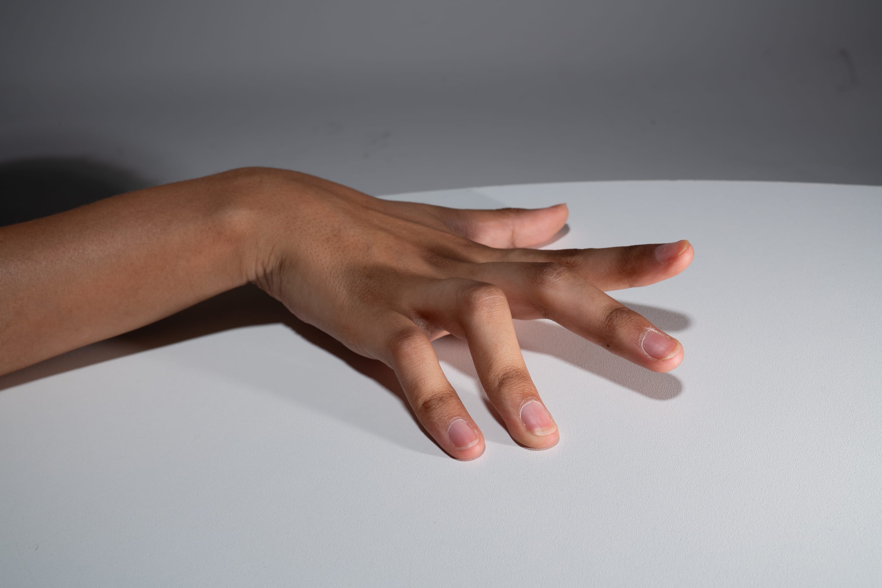 a hand with mediumtoned skin is resting on a white table. the hand is extended, with the fingers slightly curled. the image is lit with studio lighting, creating a soft shadow underneath the hand.