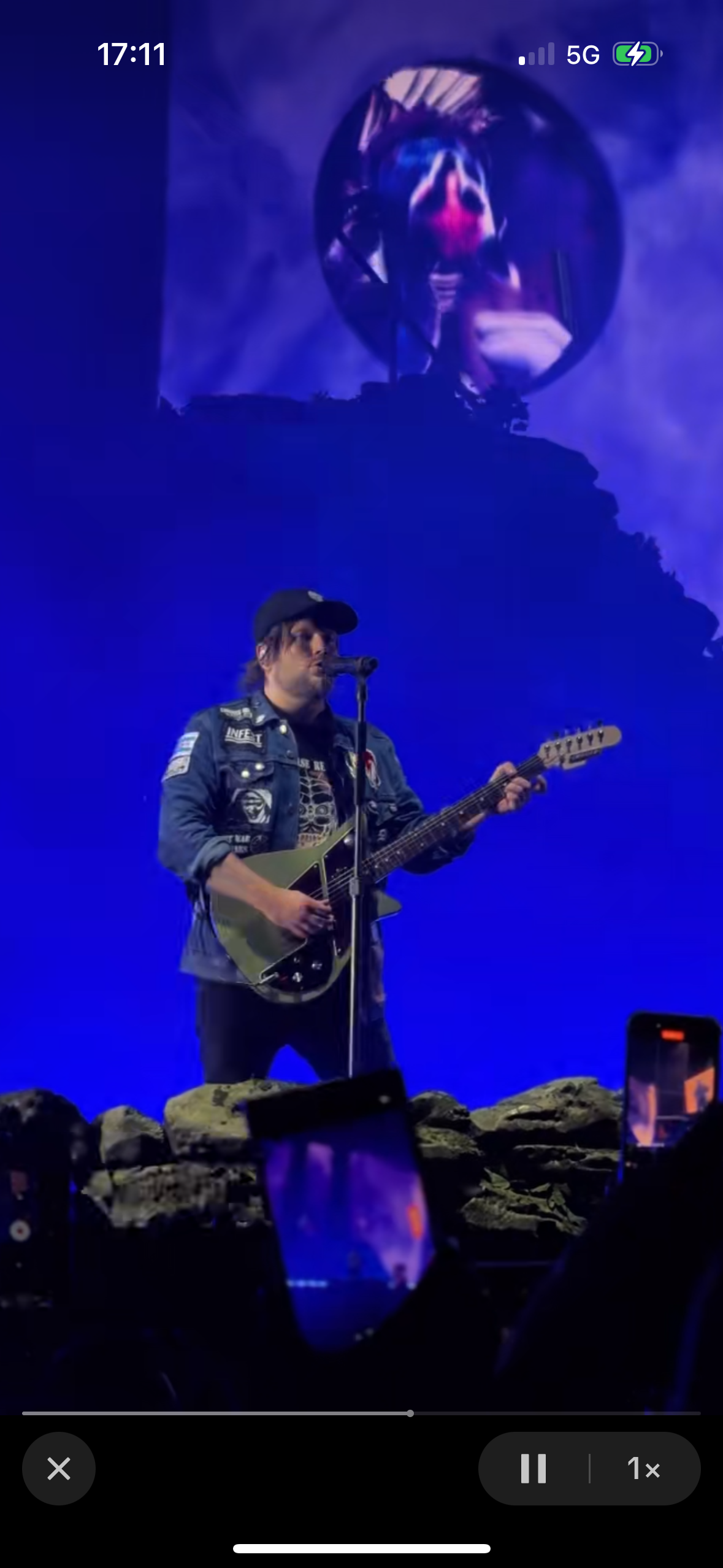 a man in a black shirt and a black cap is singing into a microphone and playing a guitar on stage. the stage is lit up with blue and purple lights.  he is performing in front of a crowd. the man is enjoying performing to his fans.