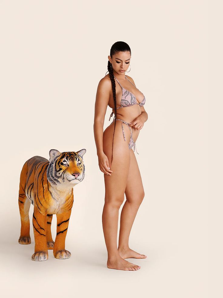 a woman with long, dark hair styled in a braid stands in a confident pose, showcasing a twopiece animal print bikini.  she is positioned beside a lifesize tiger statue against a minimalist cream backdrop. the scene is brightly lit in a professional studio setting.