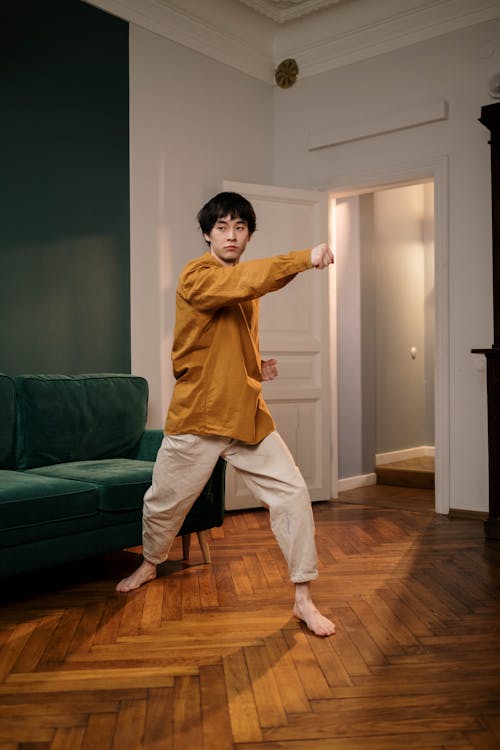 a young asian man wearing a mustardcolored shirt and beige pants practices martial arts in his living room. he's standing on a hardwood floor, striking a dynamic pose with his right arm extended. a green sofa is visible in the background. the room is stylish and modern. the overall mood is one of focused energy and selfimprovement.