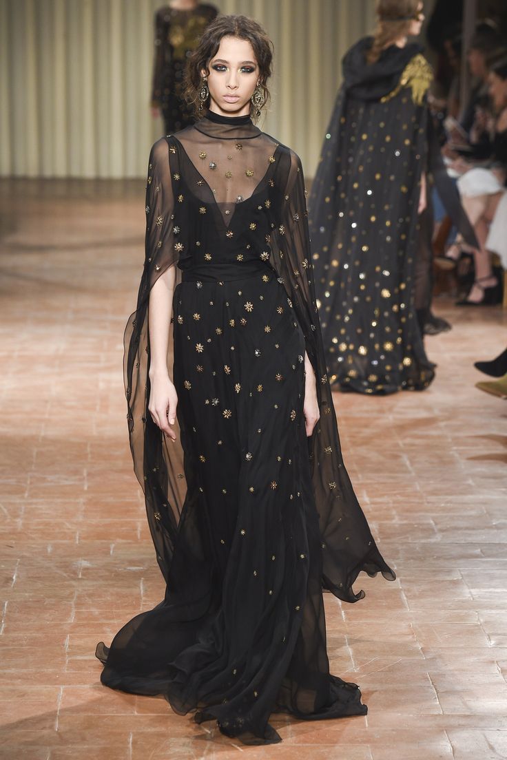 a female model walks the runway wearing a stunning black floorlength gown. the dress features a sheer overlay with gold star embellishments throughout. the sheer overlay is styled as a cape that drapes elegantly around the model. the model's hair is styled in loose curls and she wears minimal jewelry. the background is an indoor runway setting, possibly during a fashion show. the flooring appears to be a reddishbrown stone.