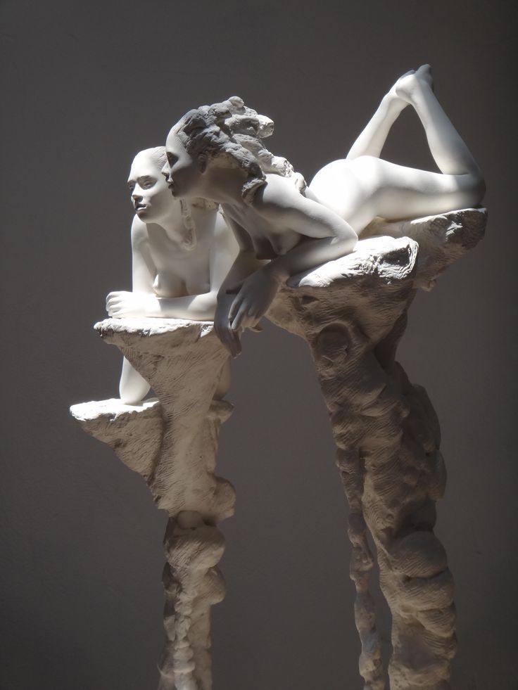 a white sculpture of two nude female figures in a classical pose. they are perched on a stylized rock formation. the lighting is dramatic, highlighting the curves of the figures and creating deep shadows. the background is a dark gray, which makes the sculpture stand out.