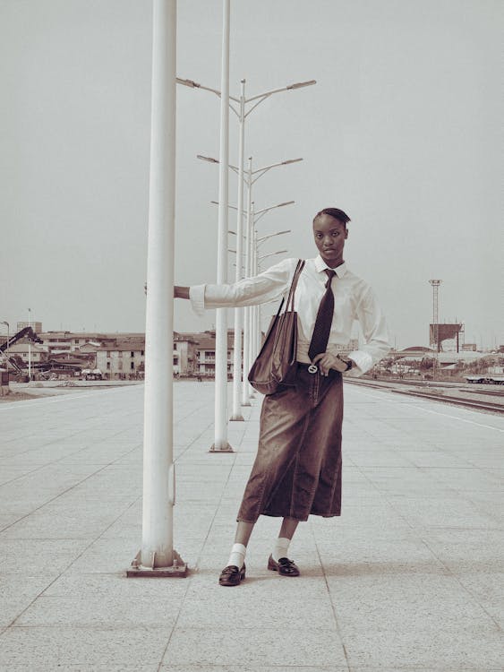 a tall, slender woman is standing on a platform, leaning on a white street light. she is wearing a white, buttonup shirt with a black tie, a long, denim skirt, white socks, and black loafers. she has a brown handbag slung over her shoulder. behind her are several street lights, a building in the distance, and train tracks. the sky is overcast.