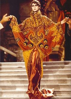 a woman in a dramatic, ornate gold and red gown stands on a runway, her pose striking and her expression confident. the gown features intricate designs and embellishments, showcasing haute couture craftsmanship. the background is blurry, suggesting the image was taken during a fashion show. the overall vibe is glamorous and elegant.