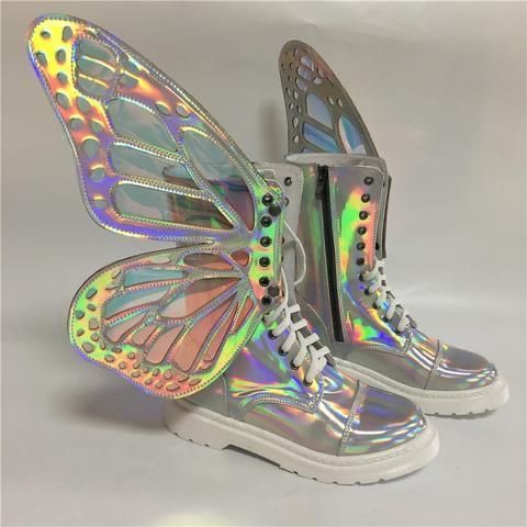 the image shows a pair of women's platform combat boots featuring large, iridescent holographic butterfly wings attached to the sides.  the boots themselves are a silveryholographic material, and have white laces and soles. one boot is slightly further back than the other, showing both boots at slightly different angles. the background is a simple, offwhite surface. the overall aesthetic is whimsical and fantastical.